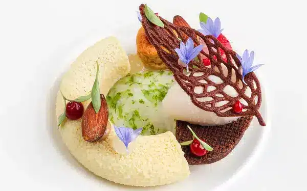 Postre artístico con base de helado o mousse, cremosos, bizcochos, flores comestibles, hojas de chocolate y frutos rojos