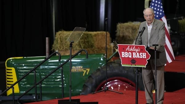 Senador Chuck Grassley en un escenario con un tractor de fondo durante un evento rural